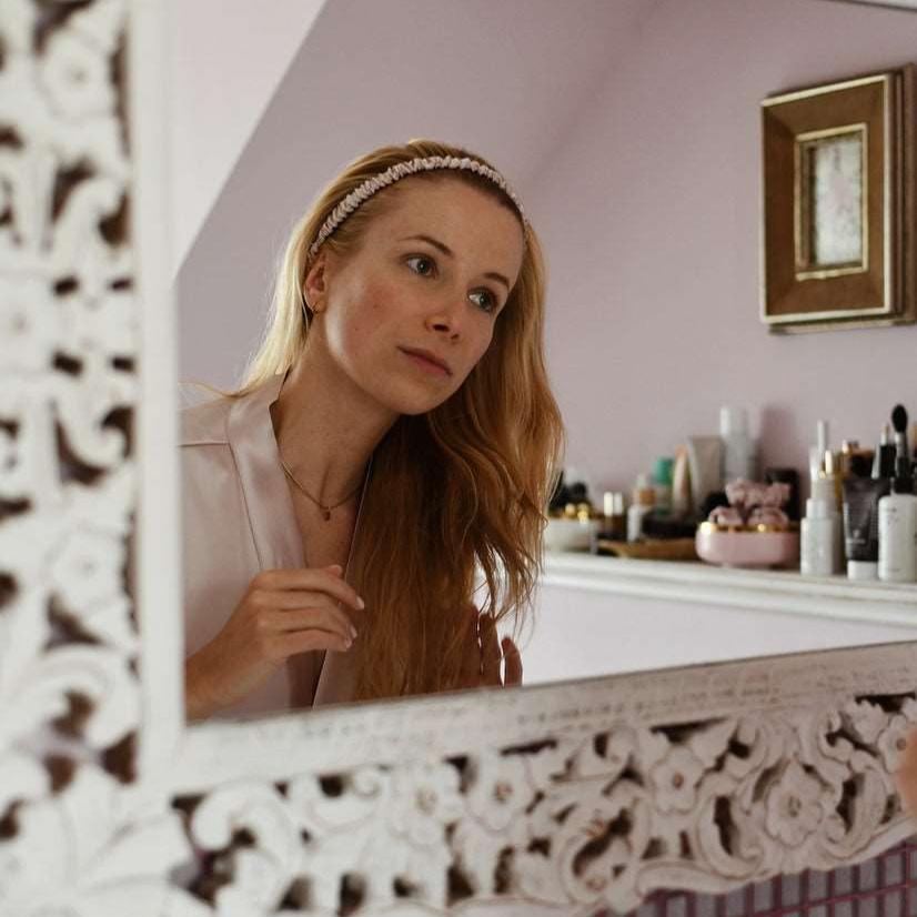 Pink Woman wearing a luxurious blush pink silk headband by Dariia Day while styling her hair in front of a mirror, perfect for gentle hair care and an elegant look.