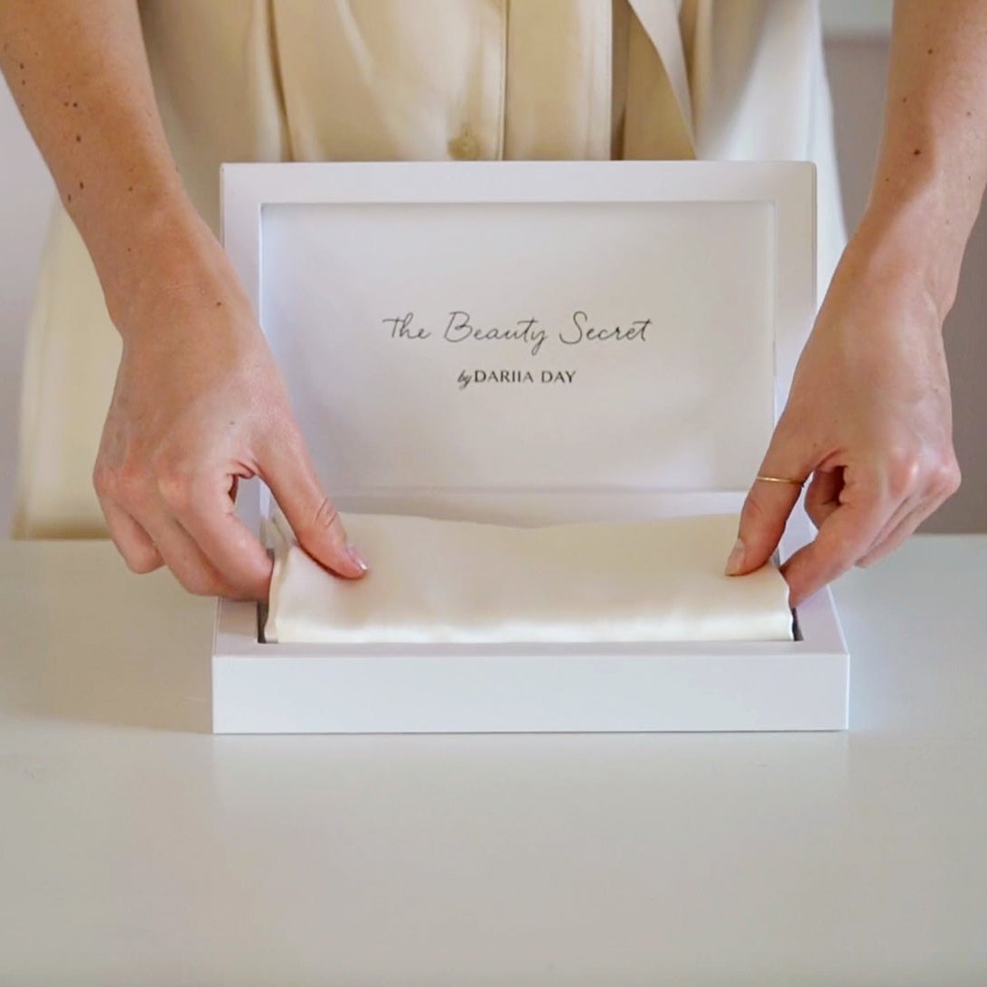 White Hands elegantly placing a Powder White mulberry silk product into a luxurious white box with the inscription 'The Beauty Secret by Dariia Day,' showcasing premium packaging and attention to detail.