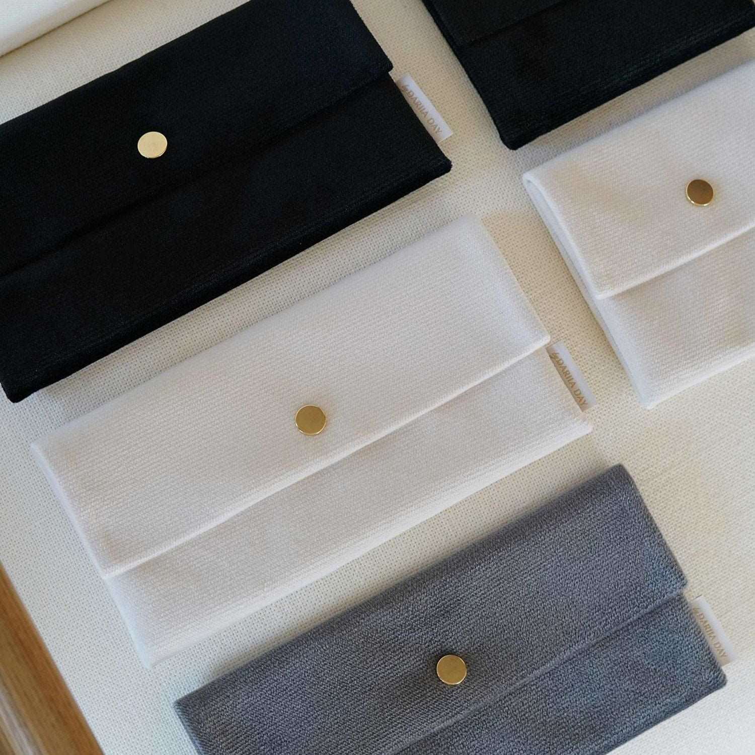 Black Dariia Day Velvet Pouch collection in Midnight Black, Powder White, and Silver Grey displayed on a beige surface.