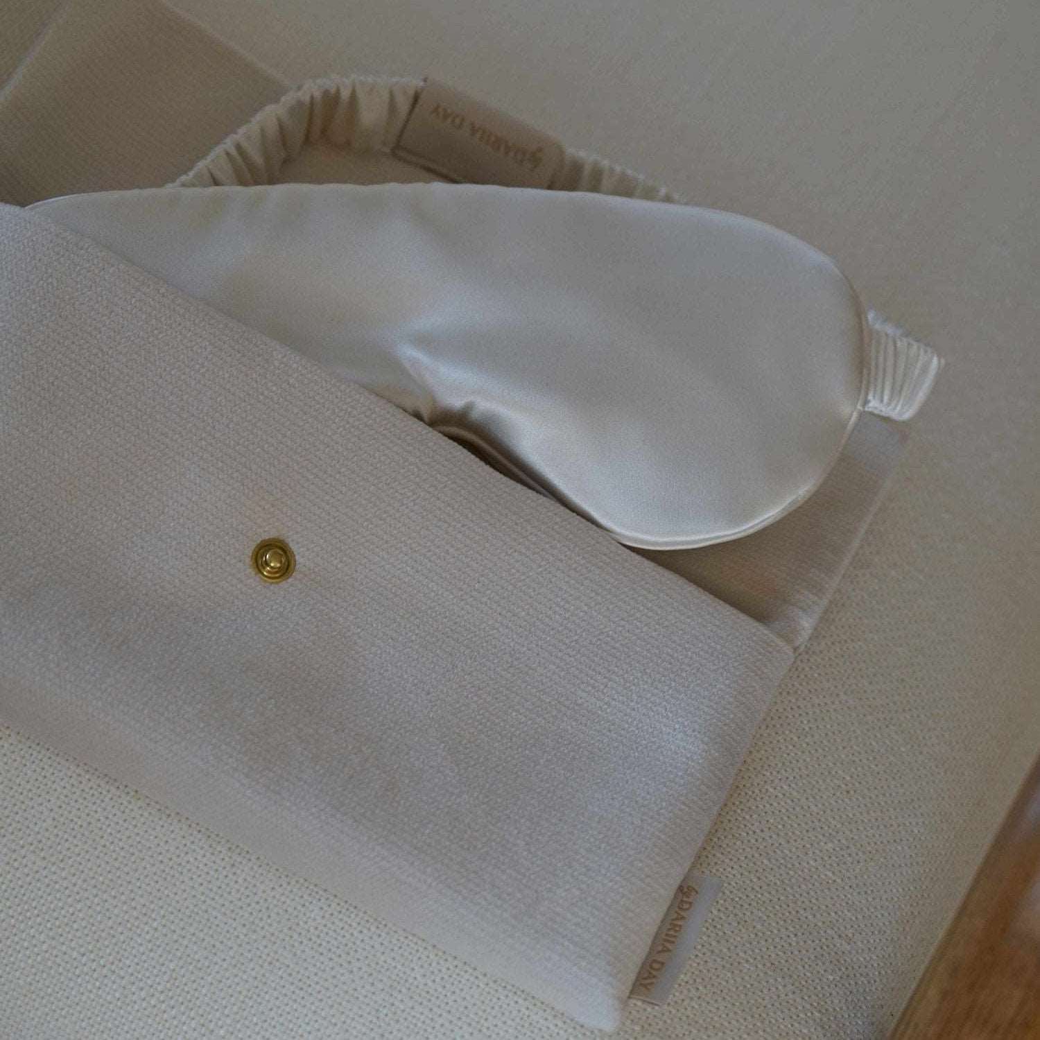 Vanilla Dariia Day White Vanilla Pouch holding a silk eye mask, displayed on a beige chair.