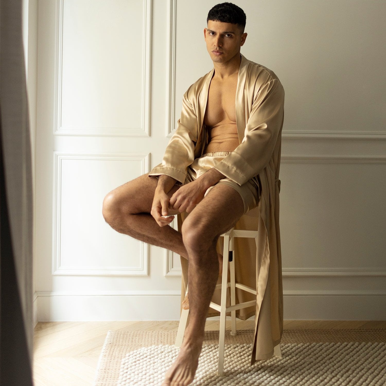 Beige  Man sitting on a stool wearing French beige silk boxers and a matching robe by Dariia Day, in a minimalistic setting.