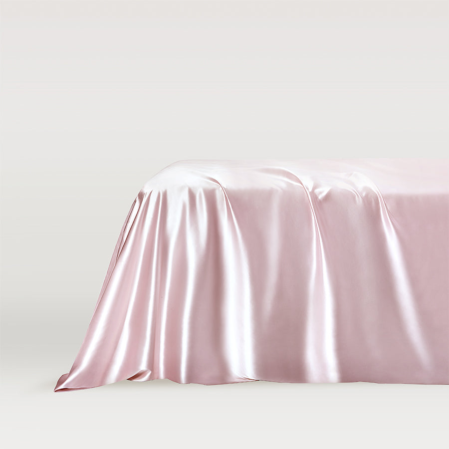 Pink Luxurious pink blush silk flat sheet draped elegantly over the corner of a bed, showcasing its smooth and glossy texture by Dariia Day.