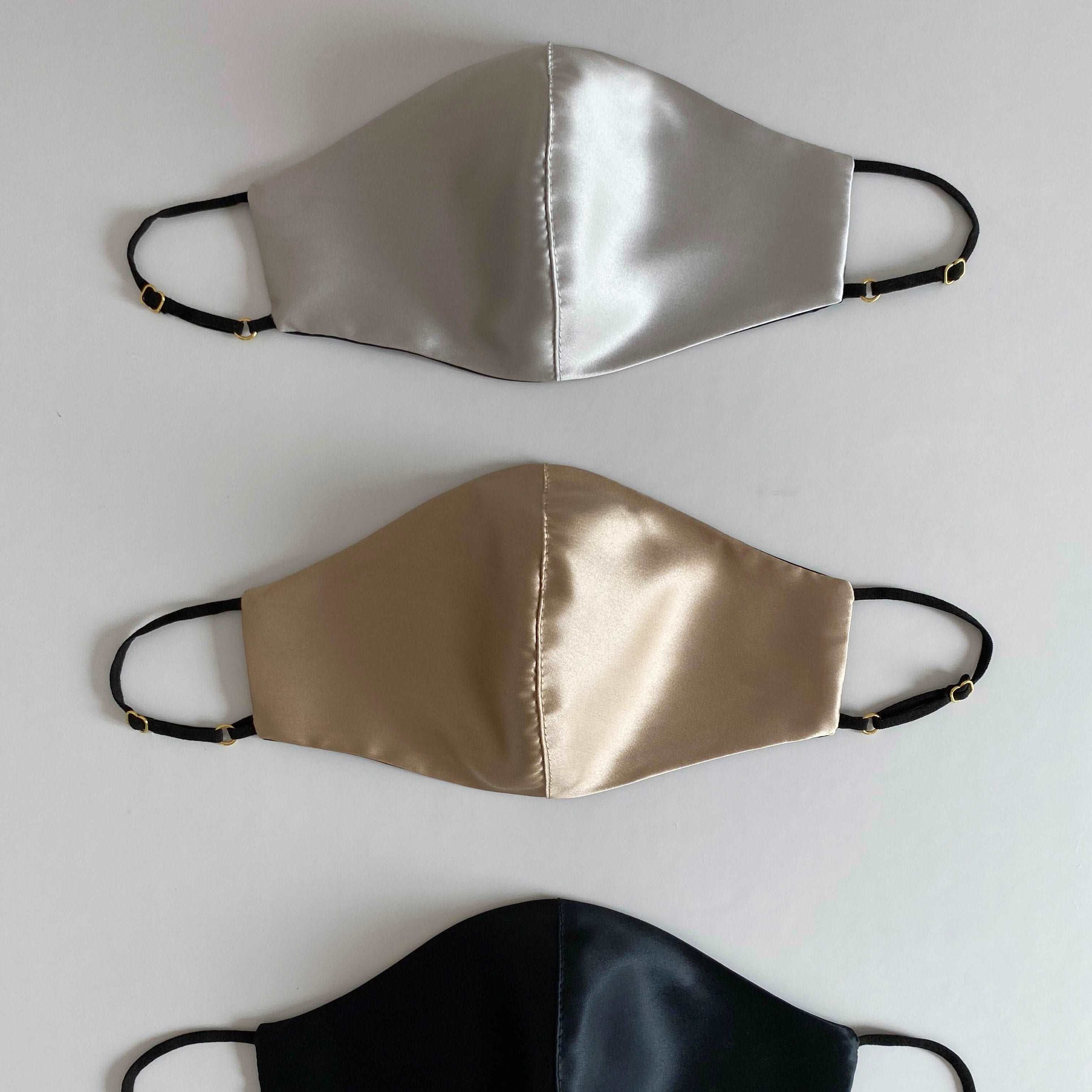 Grey Three silk face masks in French beige, silver, and black, with adjustable straps, displayed side by side against a white background