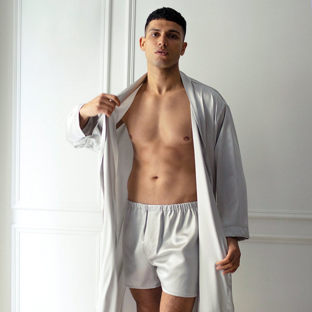 Grey Male model wearing silver grey silk boxers and an open matching robe by Dariia Day, standing in a light-filled room.