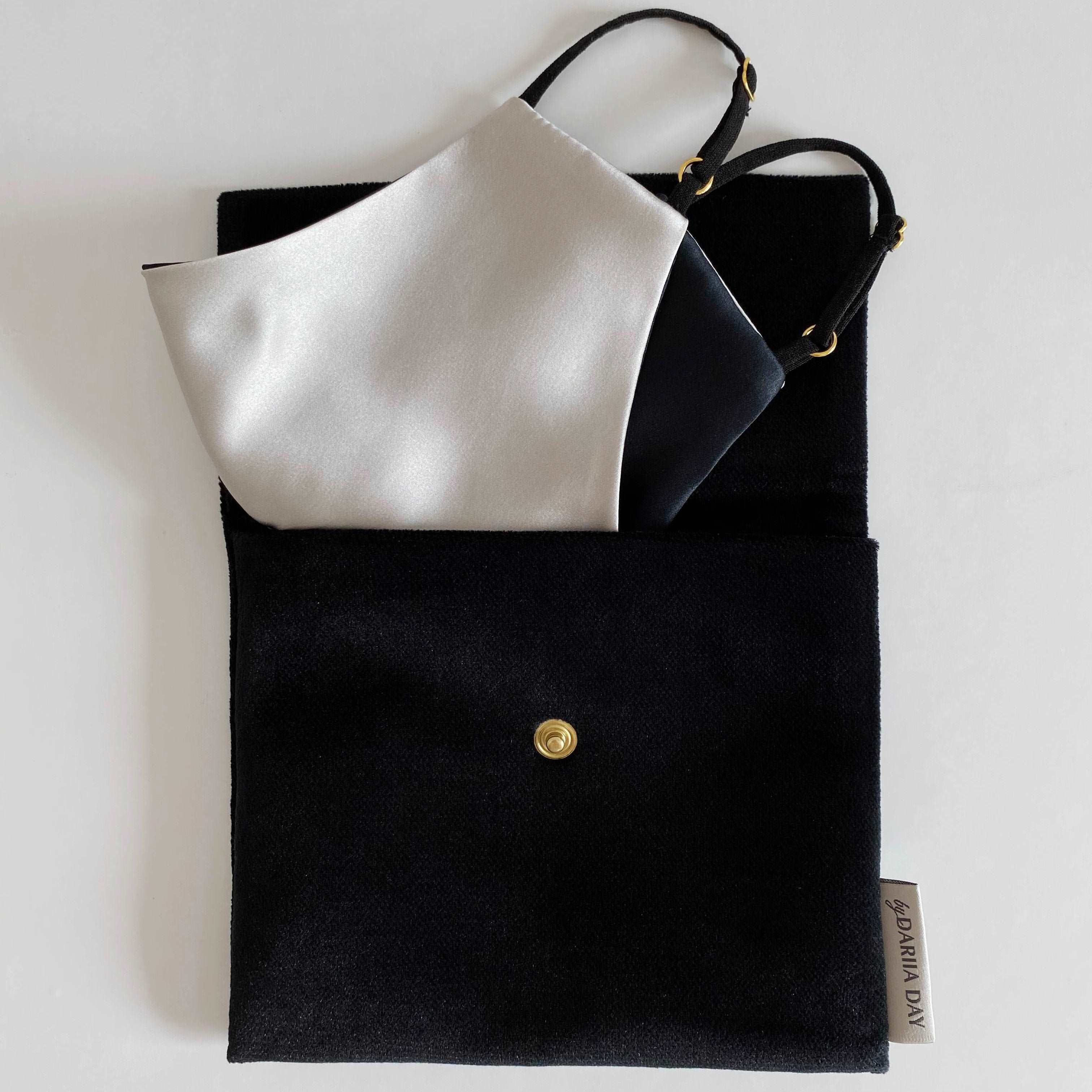 Grey Silver grey silk face mask with black adjustable straps, neatly placed inside a black fabric pouch with a gold button.