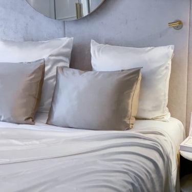 White and silver silk bedding set with a flat sheet and pillows by Dariia Day, displayed on a stylish bed with a bedside table, exuding a luxurious and comfortable vibe.