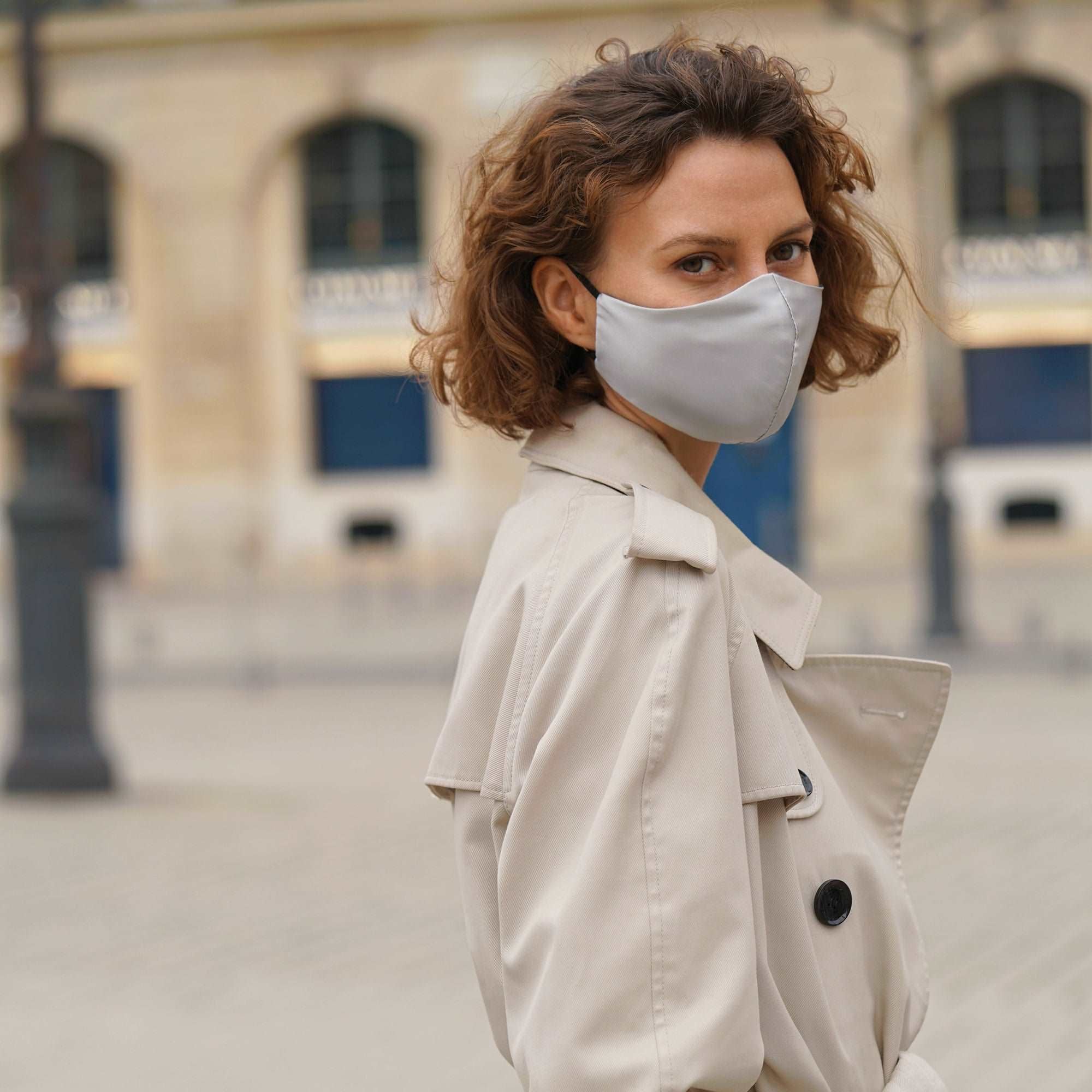 Grey Fashionable woman wearing a silver grey silk face mask with a beige trench coat, posing in an urban setting.