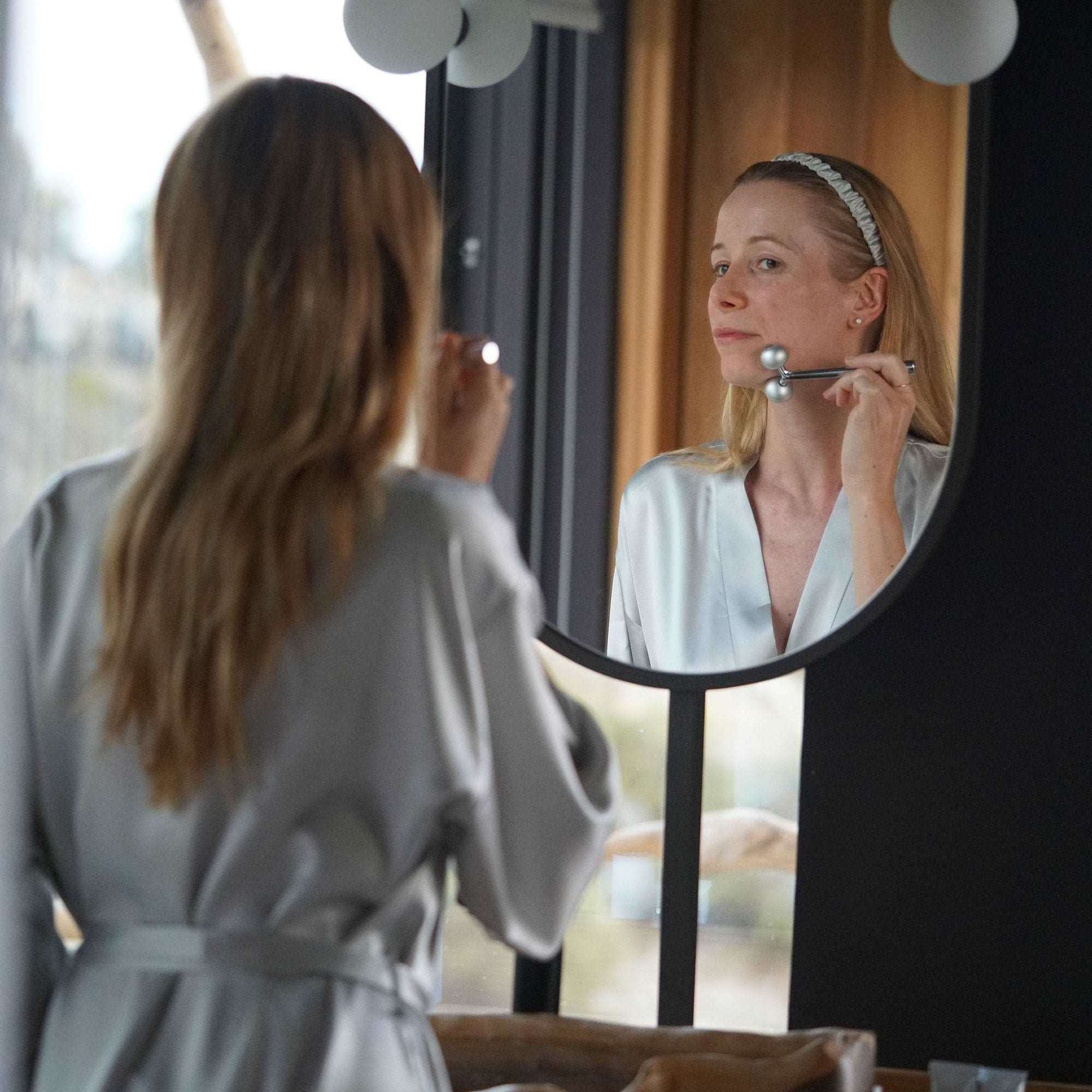 Grey Woman performing her morning skincare routine while wearing a silver grey silk robe by Dariia Day in a luxurious bathroom setting.