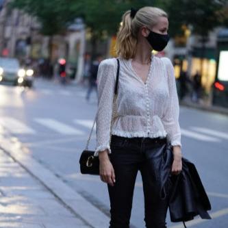 Black Fashionable woman in a white blouse and black leather pants wearing a midnight black silk face mask while standing on a city street.