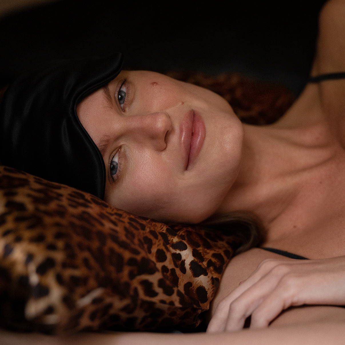 Leopard Close-up of a relaxed woman lying on a leopard-print silk pillow, wearing a black silk eye mask, highlighting the elegance of the Daria Day Leopard collection."