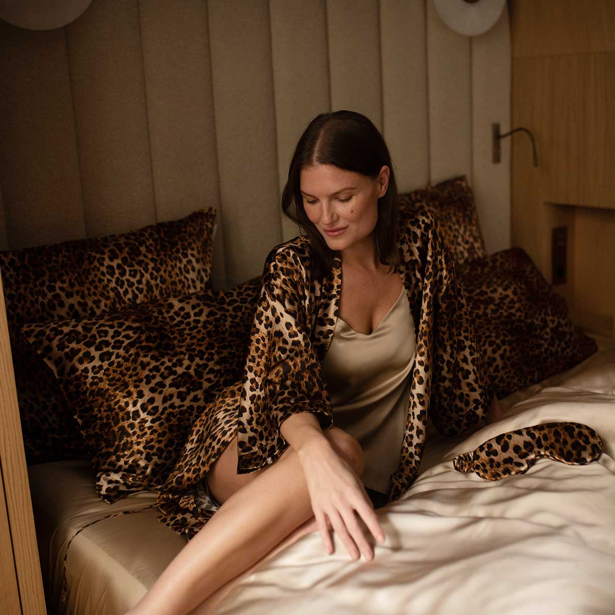 Graceful woman sitting on a bed with leopard-print silk bedding, dressed in a matching leopard-print robe and golden slip from the Daria Day Leopard collection.