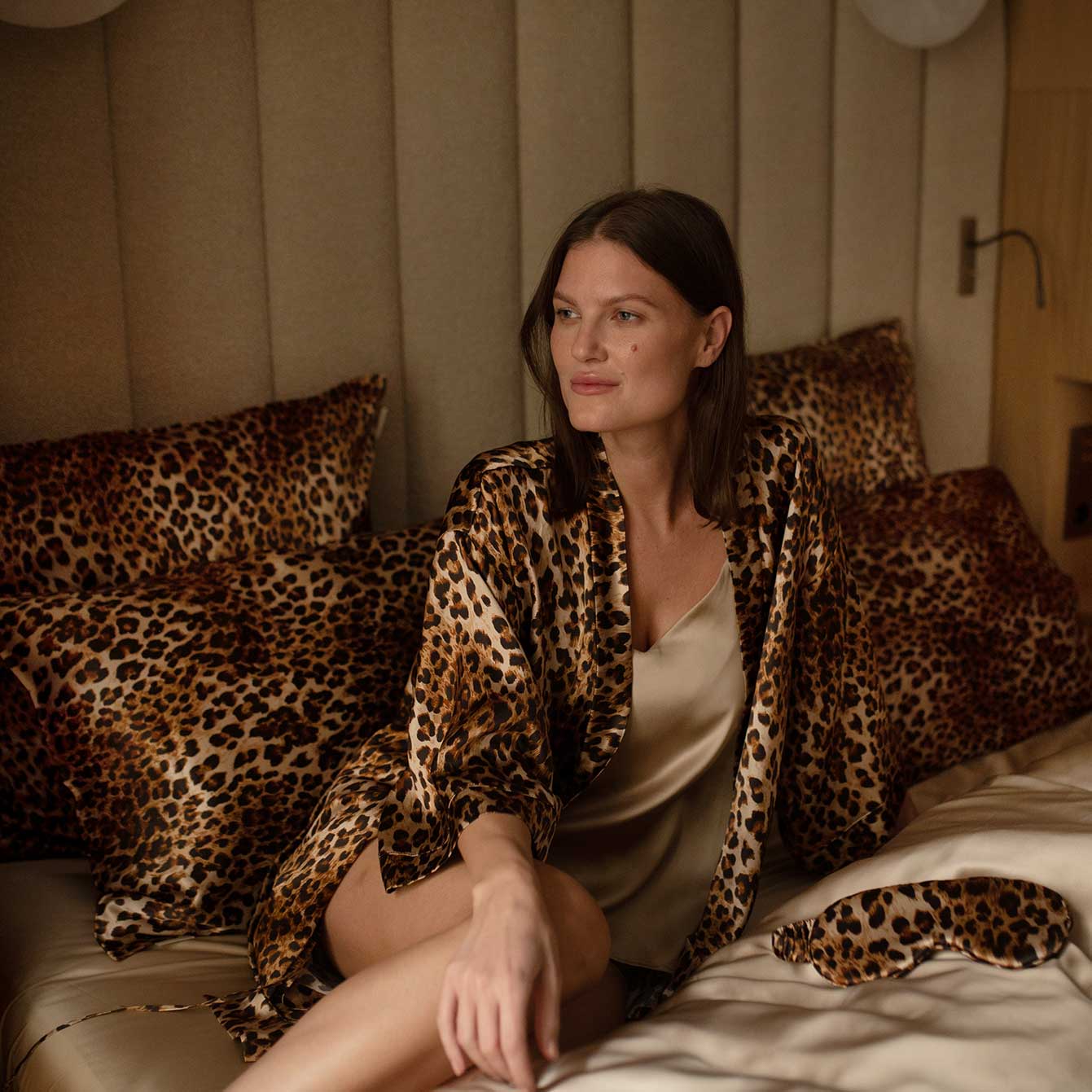 Elegant woman sitting on a bed adorned with leopard-print silk pillows, wearing a matching leopard-print silk robe from the Daria Day Leopard collection.