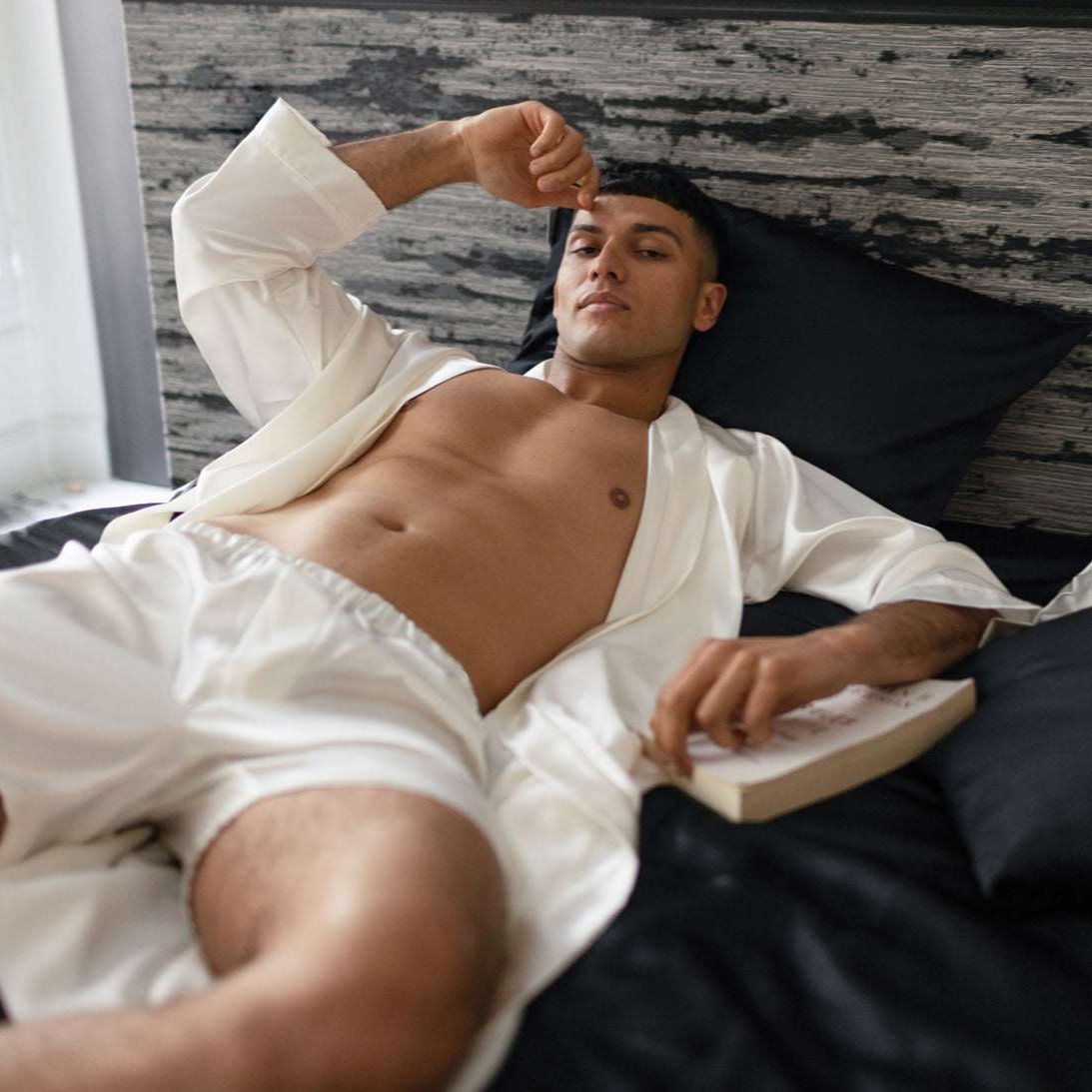 Man lounging on a bed wearing an elegant ivory silk robe and shorts by Dariia Day, surrounded by luxurious black and white silk bedding.