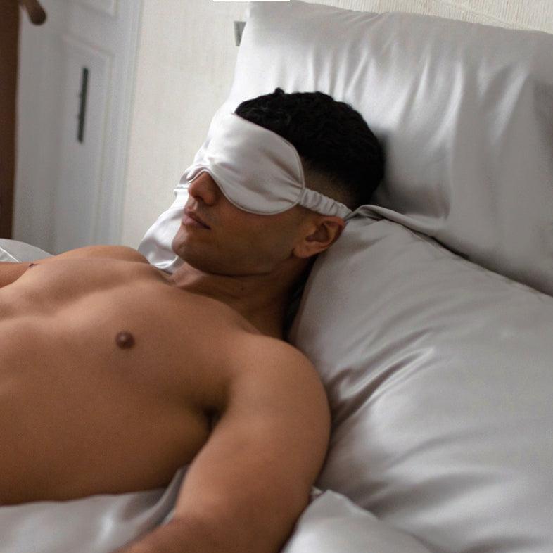 Man sleeping on luxurious silver grey silk bedding, wearing a silk eye mask from by Dariia Day.