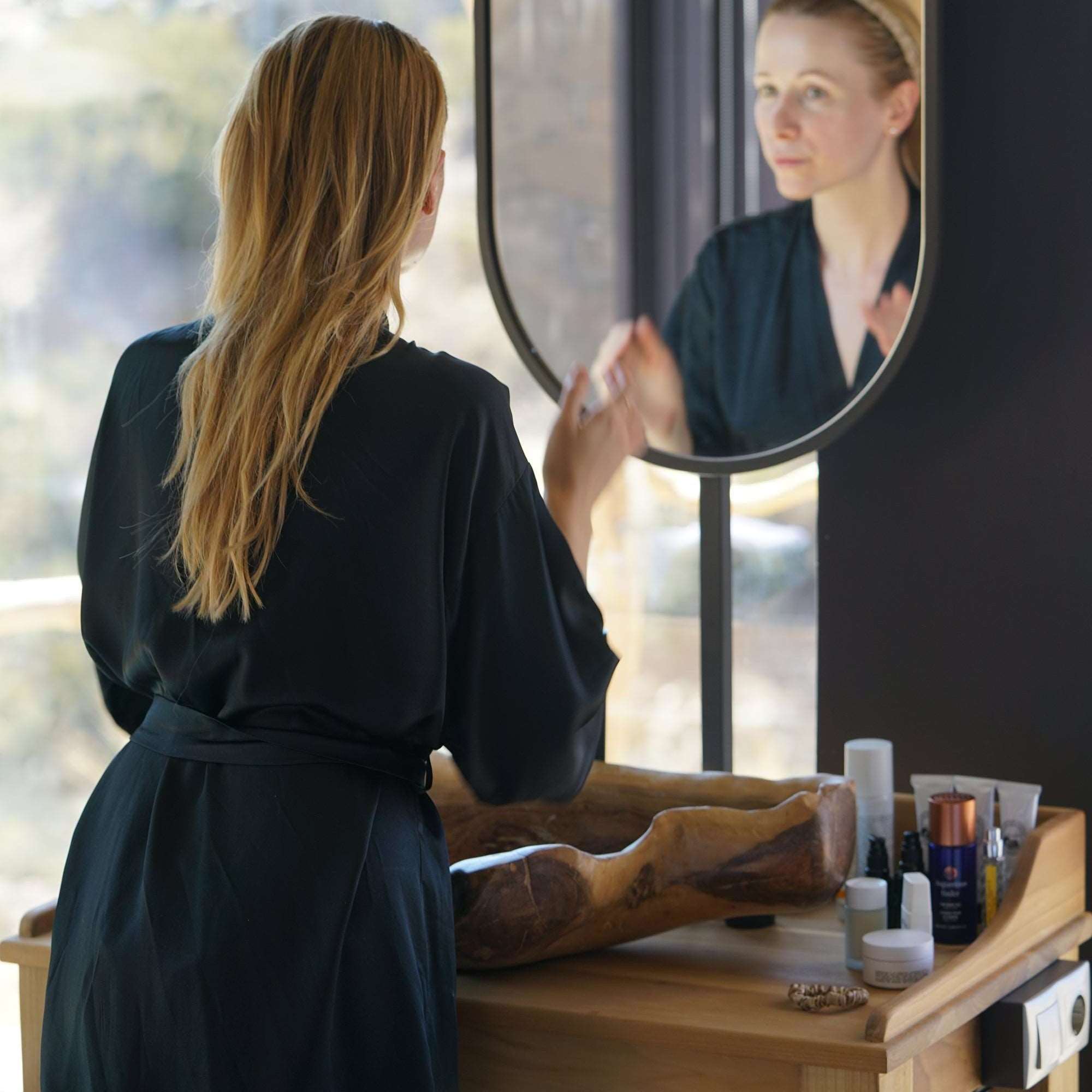 A woman in a black silk robe standing in front of a vanity mirror, applying her skincare products with a scenic view in the background.