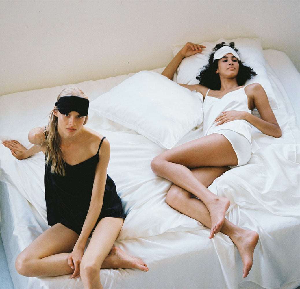 Two models lounging in silk sleepwear on a silk bed, one in black and one in white, capturing luxurious relaxation by Dariia Day.