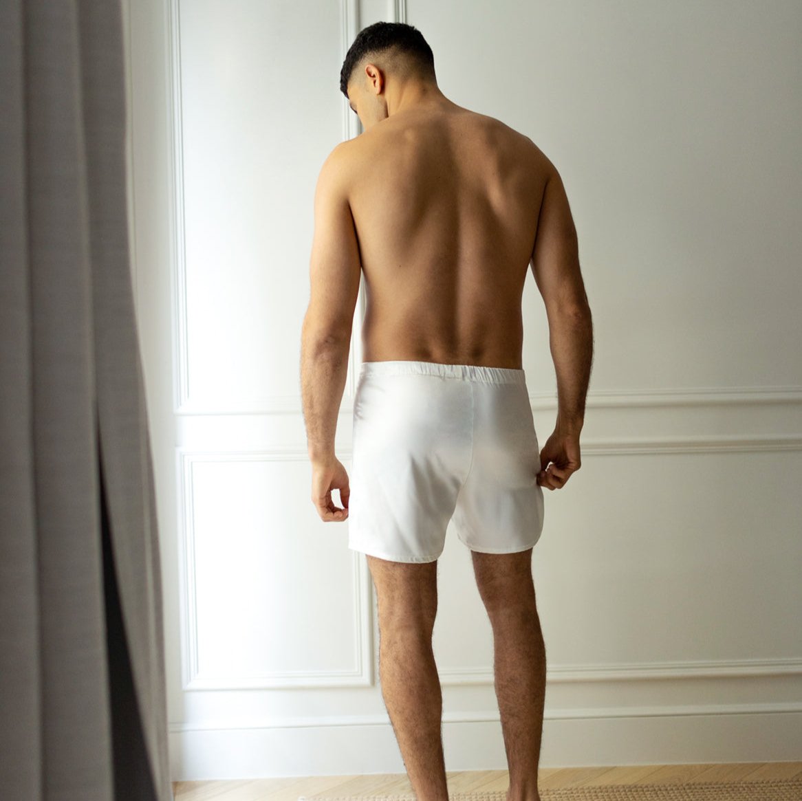 Rear view of a man in white silk boxers by Dariia Day, standing in a minimalist room.
