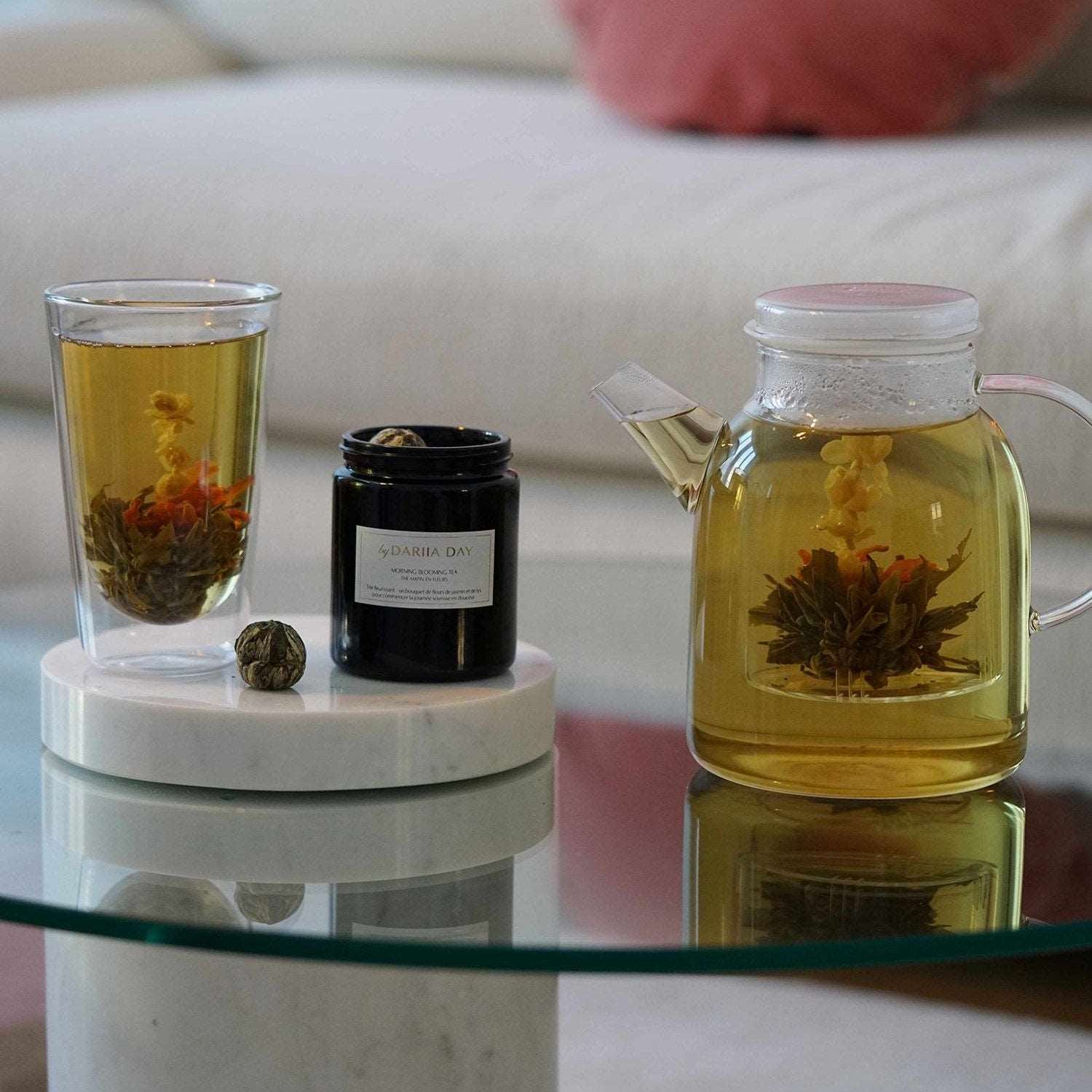 Dariia Day Morning Blooming Tea in a glass with blooming flowers, accompanied by a Silky Body Candle on a marble coaster.