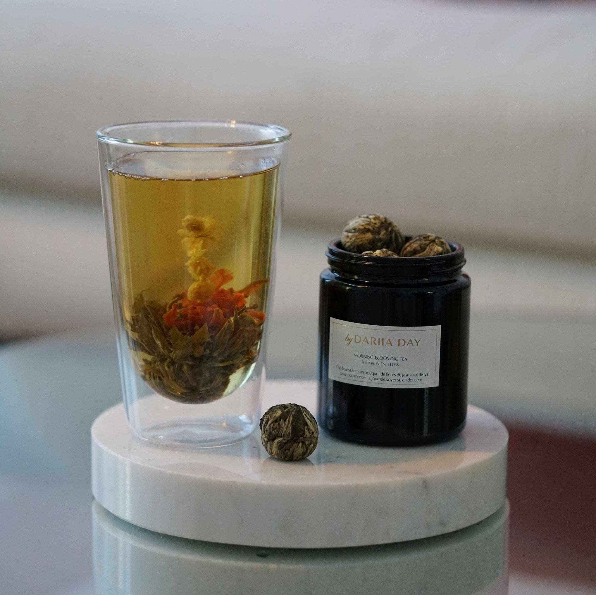 Glass of Morning Blooming Tea with blooming flowers and jar of tea by Dariia Day, placed on a marble coaster.