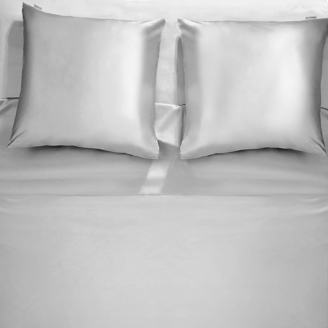 A complete silver grey silk bedding set by Dariia Day, including two pillowcases and a smooth silk sheet on a bed.