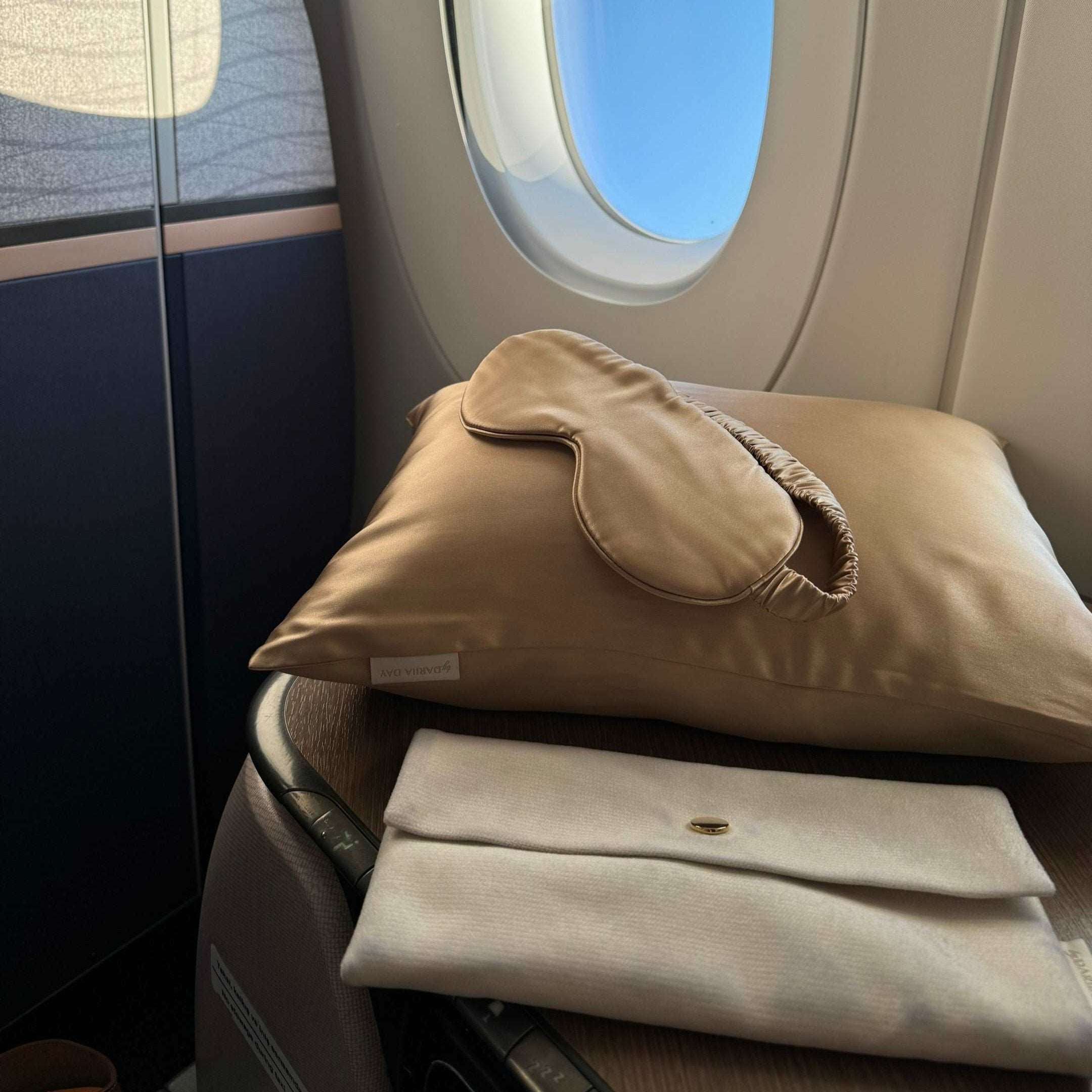 Dariia Day French beige silk travel pillow with matching eye mask on an airplane seat, next to a window.