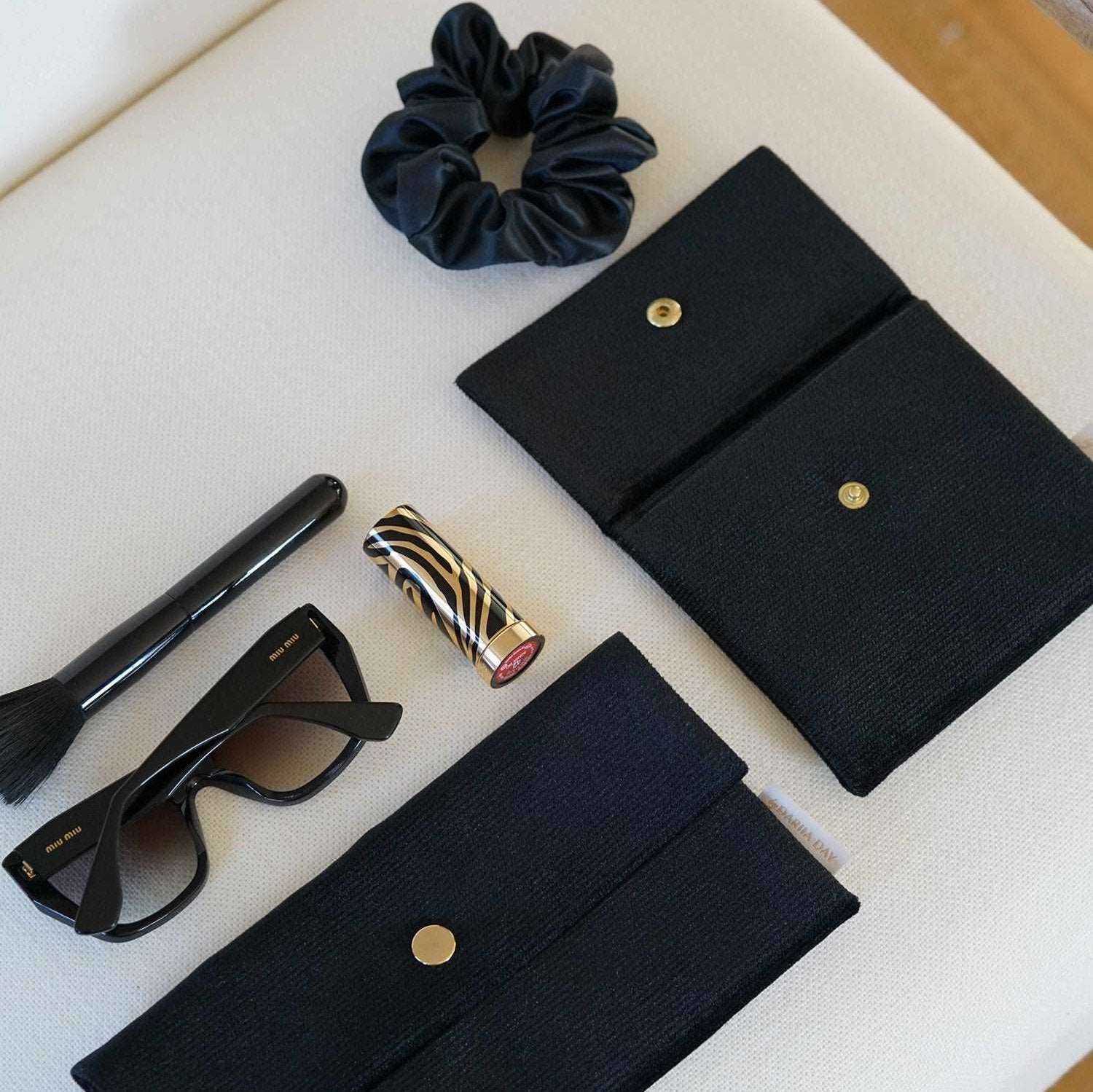 Dariia Day Midnight Black Velvet Pouch with sunglasses, lipstick, and other accessories, displayed on a beige surface