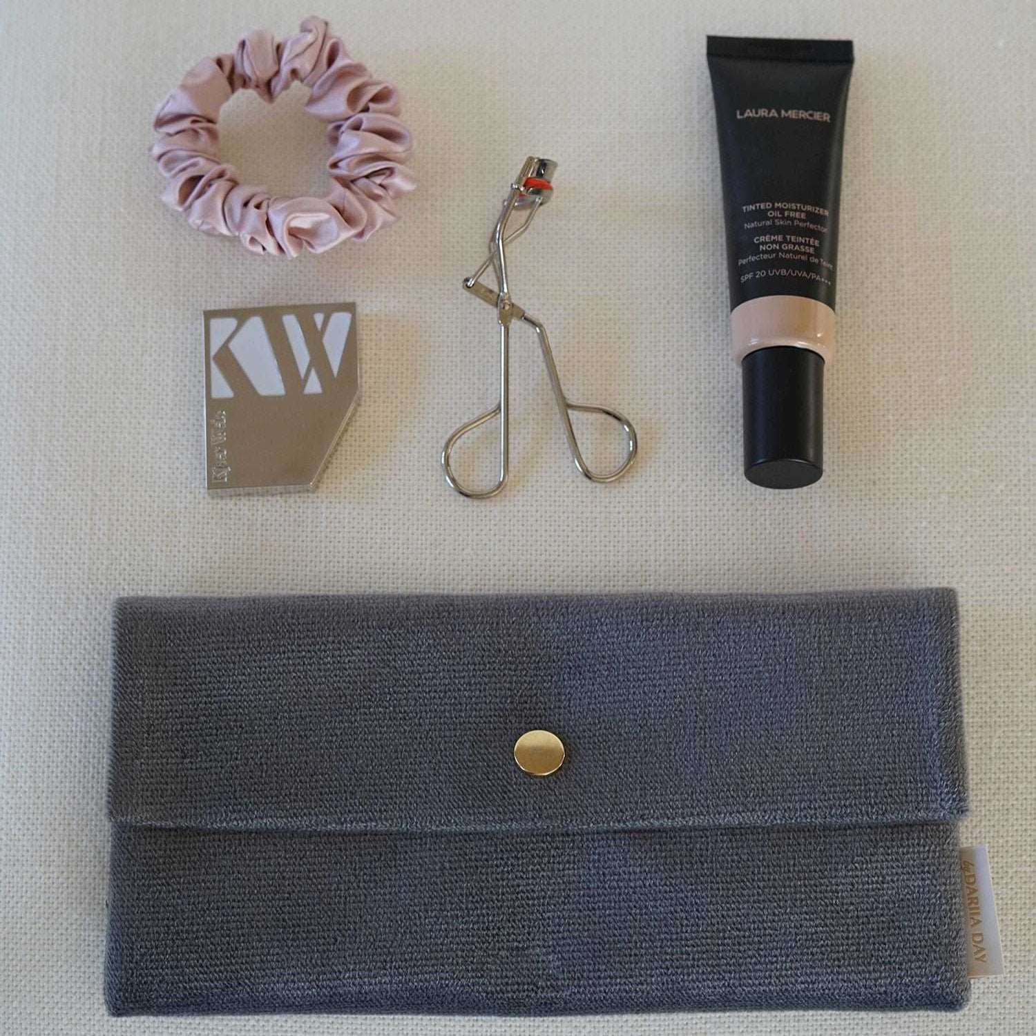 Flatlay of Dariia Day Moonstone Velvet Pouch with beauty accessories, including a pink silk scrunchie, eyelash curler, and makeup items.