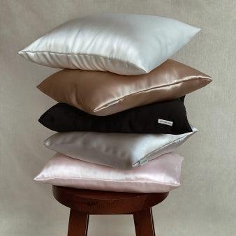 Assorted silk pillows stacked on a wooden stool by Dariia Day, featuring colors like white, beige, black, silver, and pink.