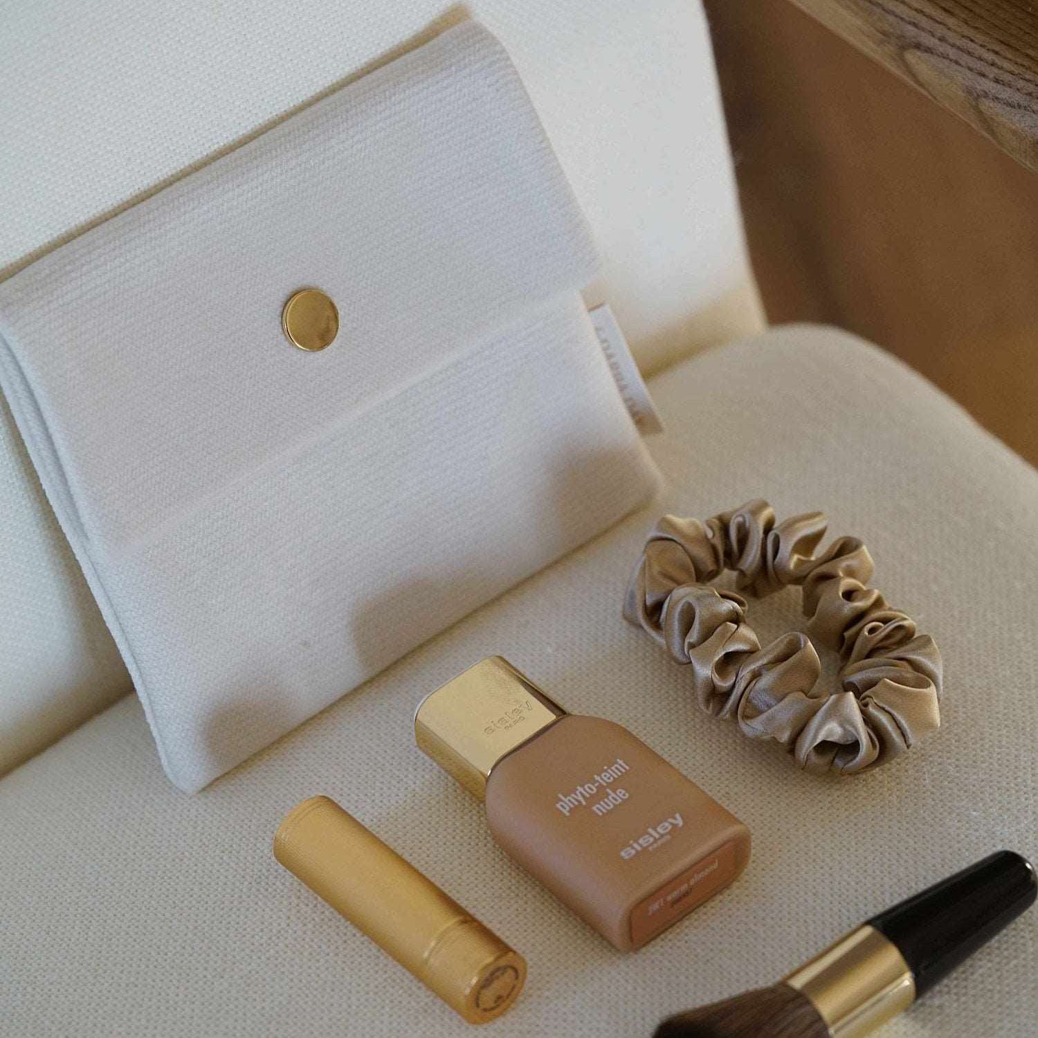 White Vanilla Velvet Pouch by Dariia Day displayed with beige scrunchie, Sisley foundation, gold lipstick, and makeup brush on a beige chair.