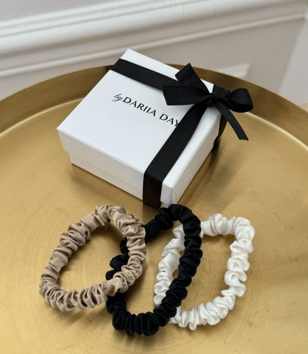 Set of 3 XS silk scrunchies in French Beige, Midnight Black, and Powder White by Dariia Day, placed in front of a gift box with a black ribbon, on a gold tray.