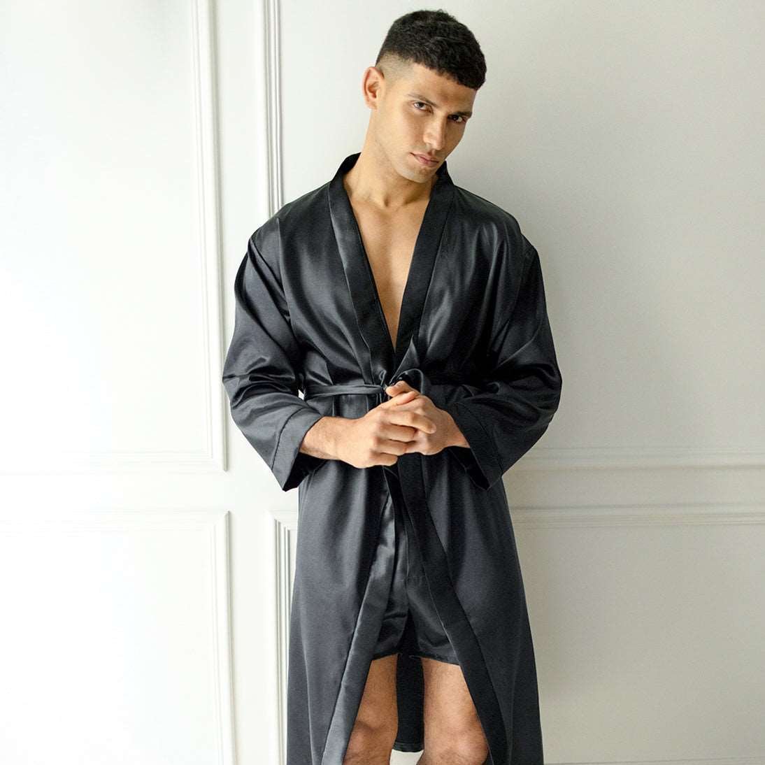 Man wearing a black silk robe with matching shorts by Dariia Day, standing against a wall.