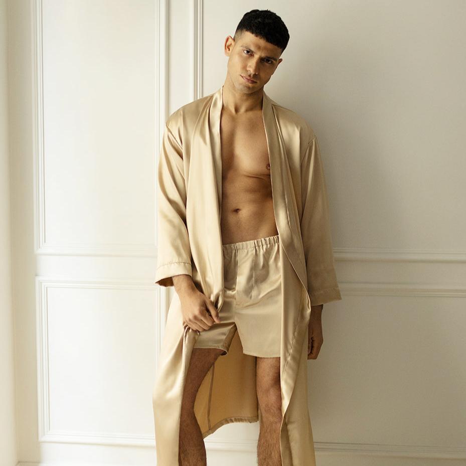 Man wearing French beige silk boxers and a matching robe by Dariia Day, standing against a white wall.
