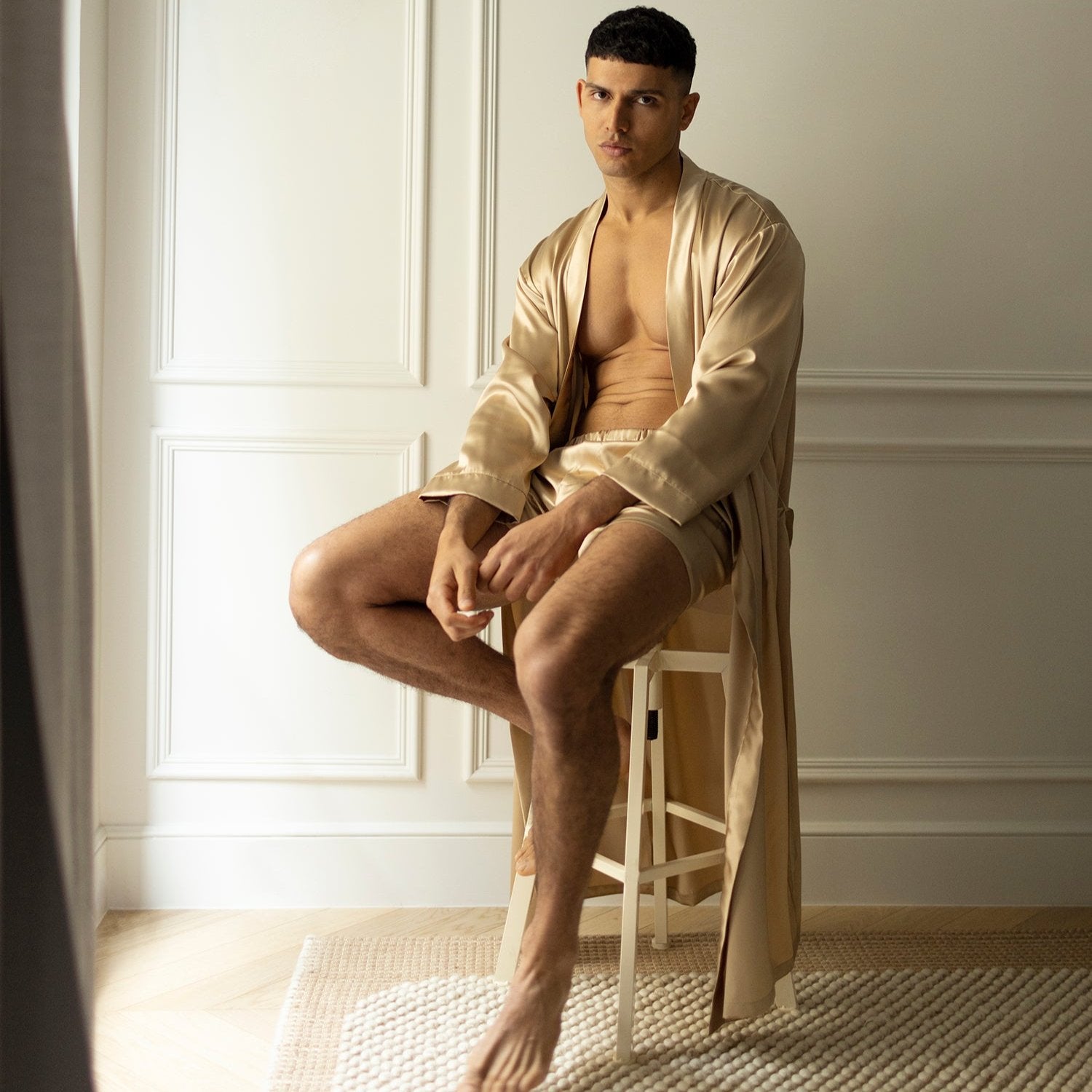  Man sitting on a stool wearing French beige silk boxers and a matching robe by Dariia Day, in a minimalistic setting.
