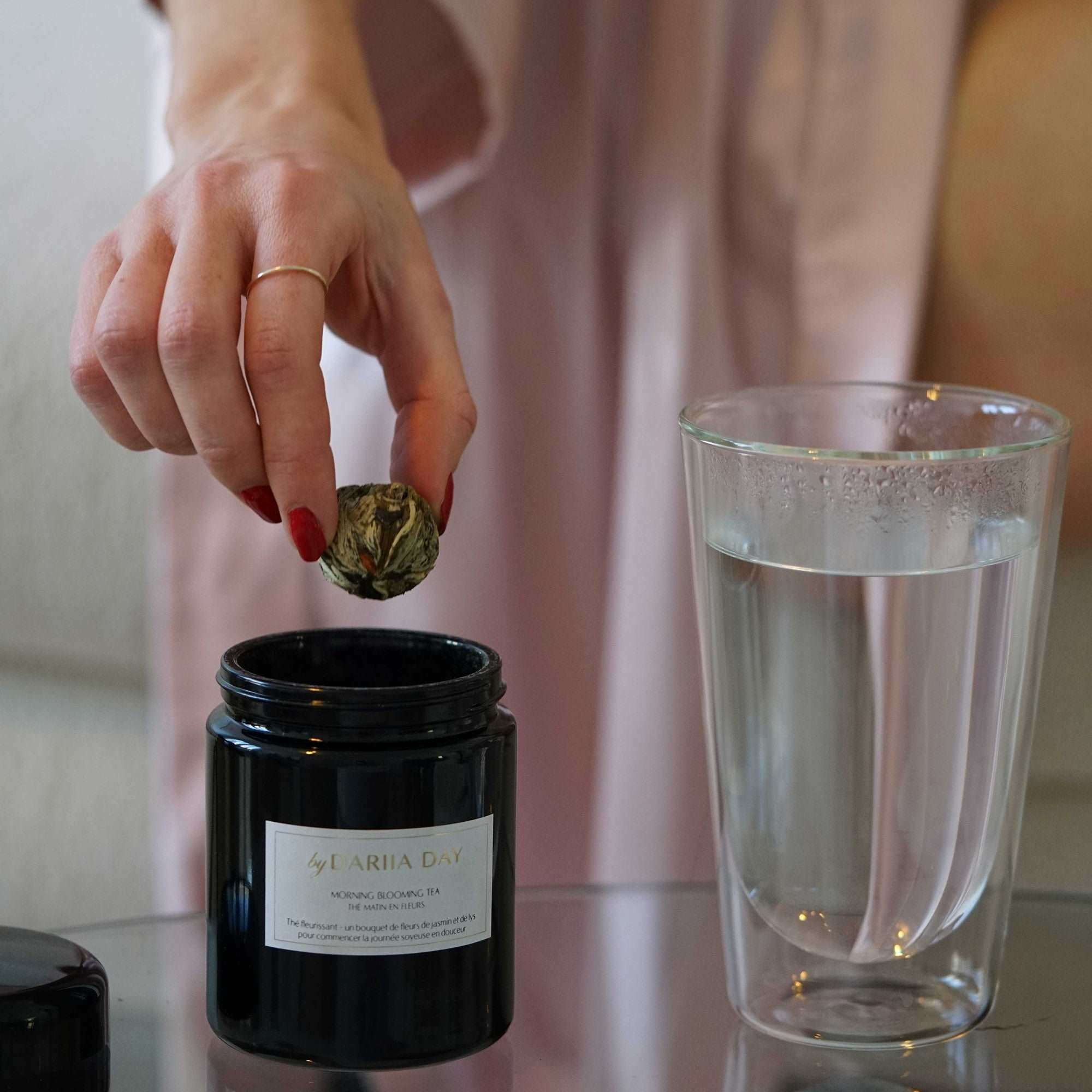 Hand dropping a blooming tea ball into a glass, preparing Morning Blooming Tea by Dariia Day.