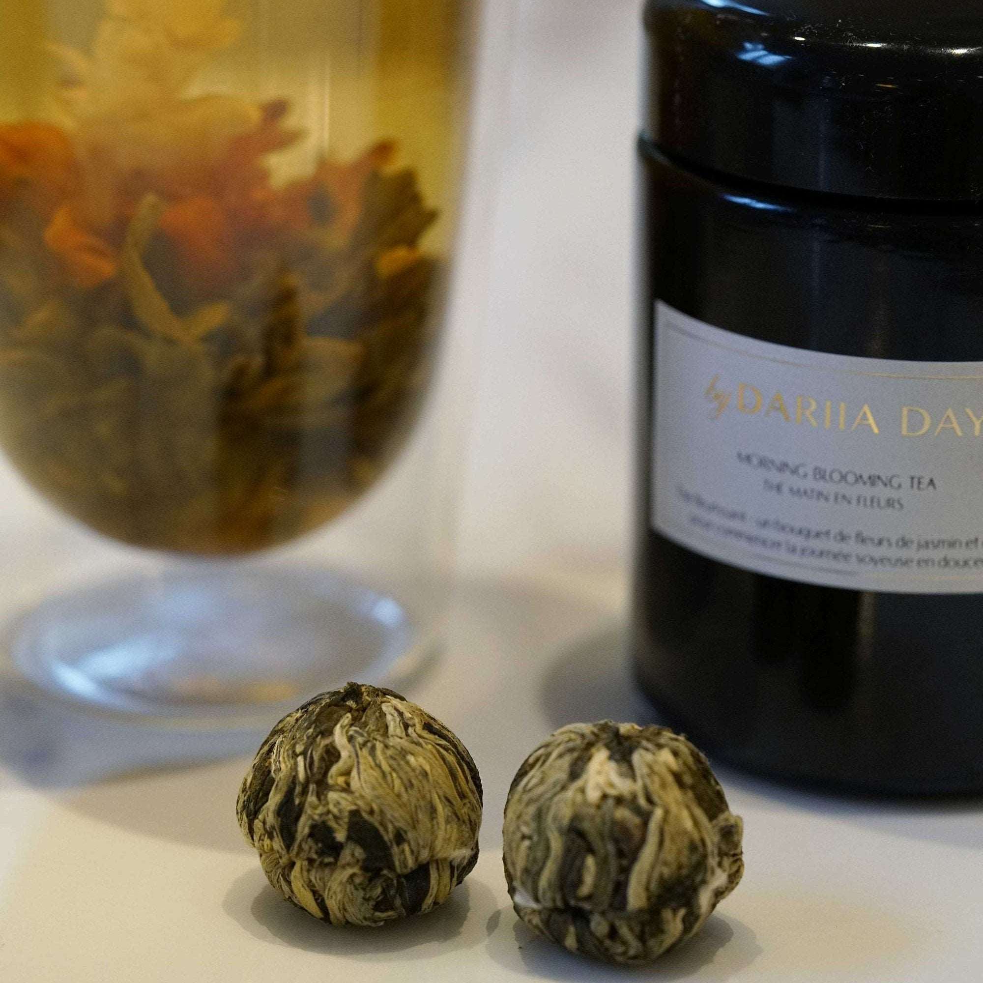 Close-up of two blooming tea balls with a glass of tea and jar of Morning Blooming Tea by Dariia Day in the background.