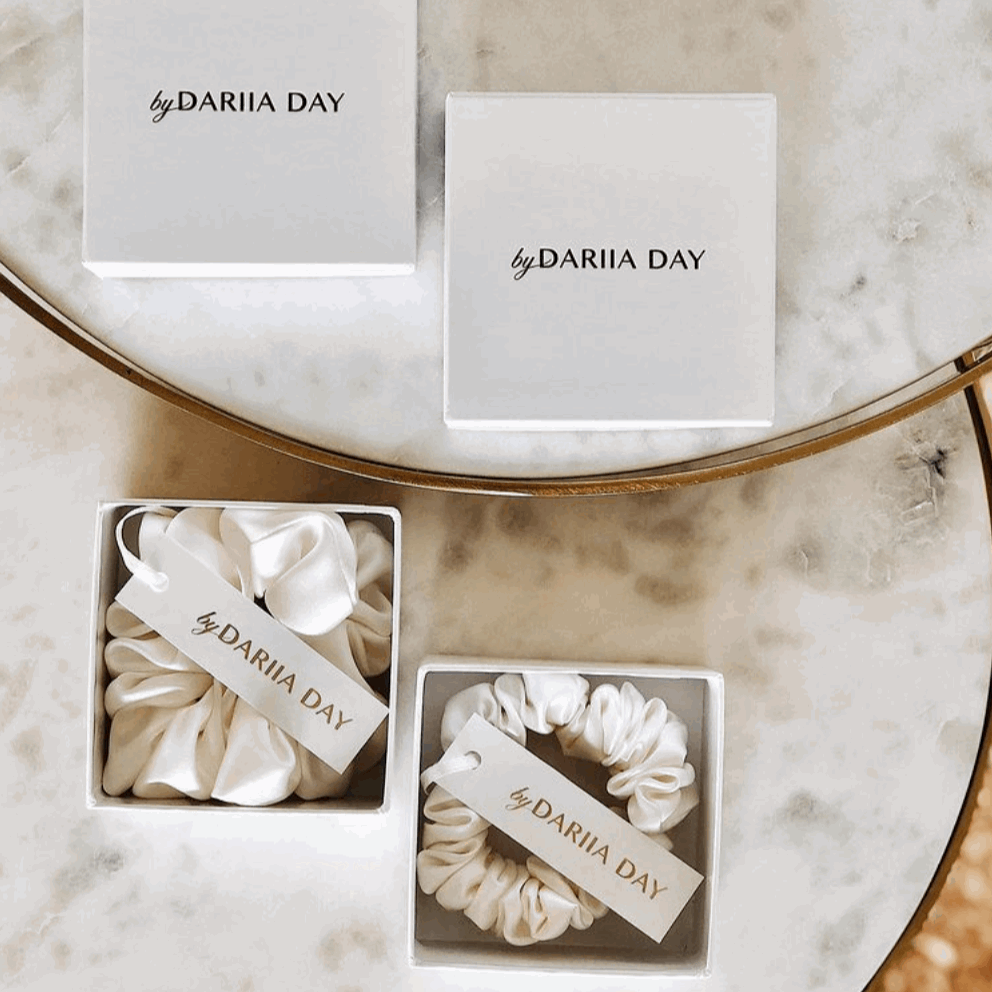 Two powder white silk scrunchies by Dariia Day displayed in elegant gift boxes, a luxurious accessory for hair.