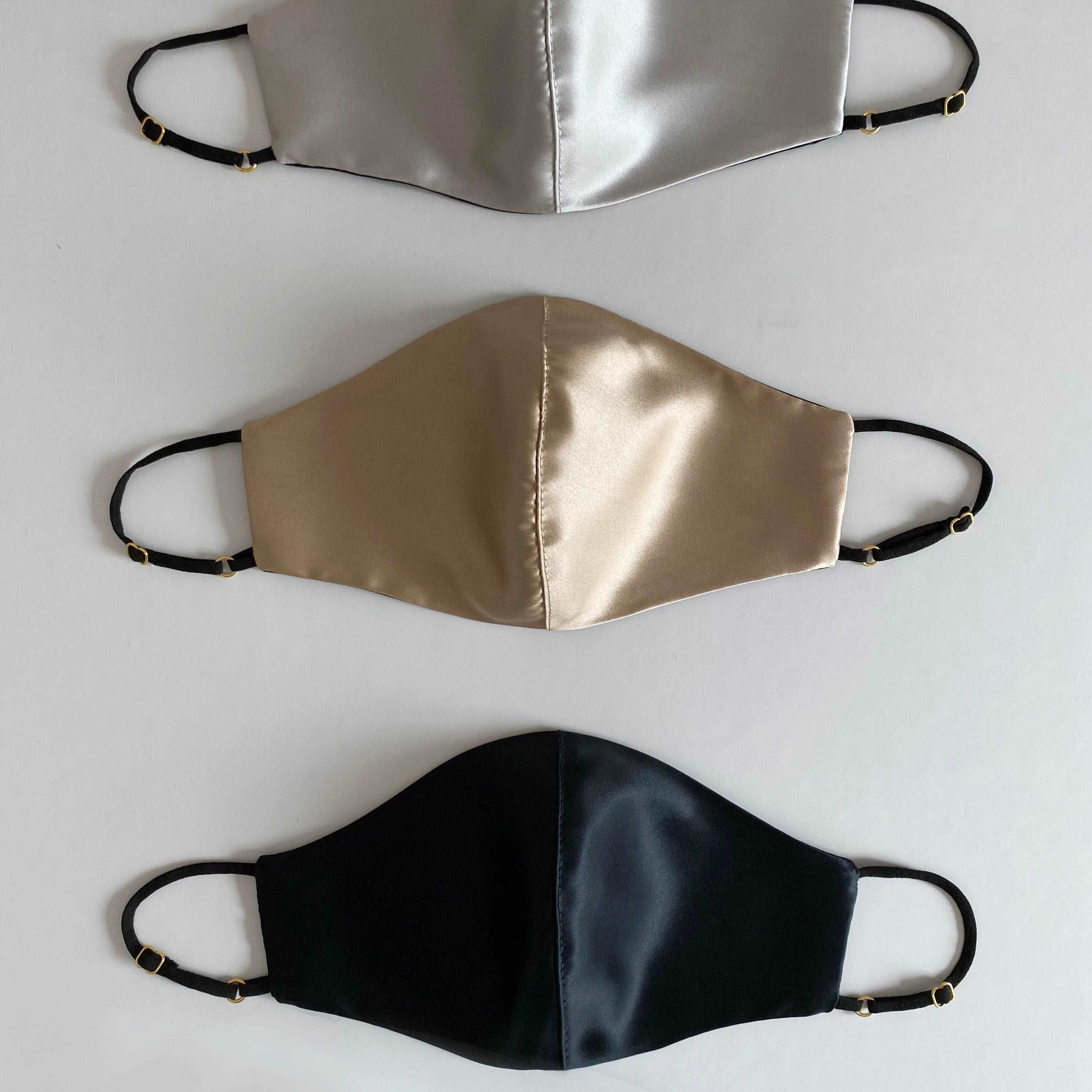 Three silk face masks in French beige, silver, and black, with adjustable straps, displayed side by side against a white background