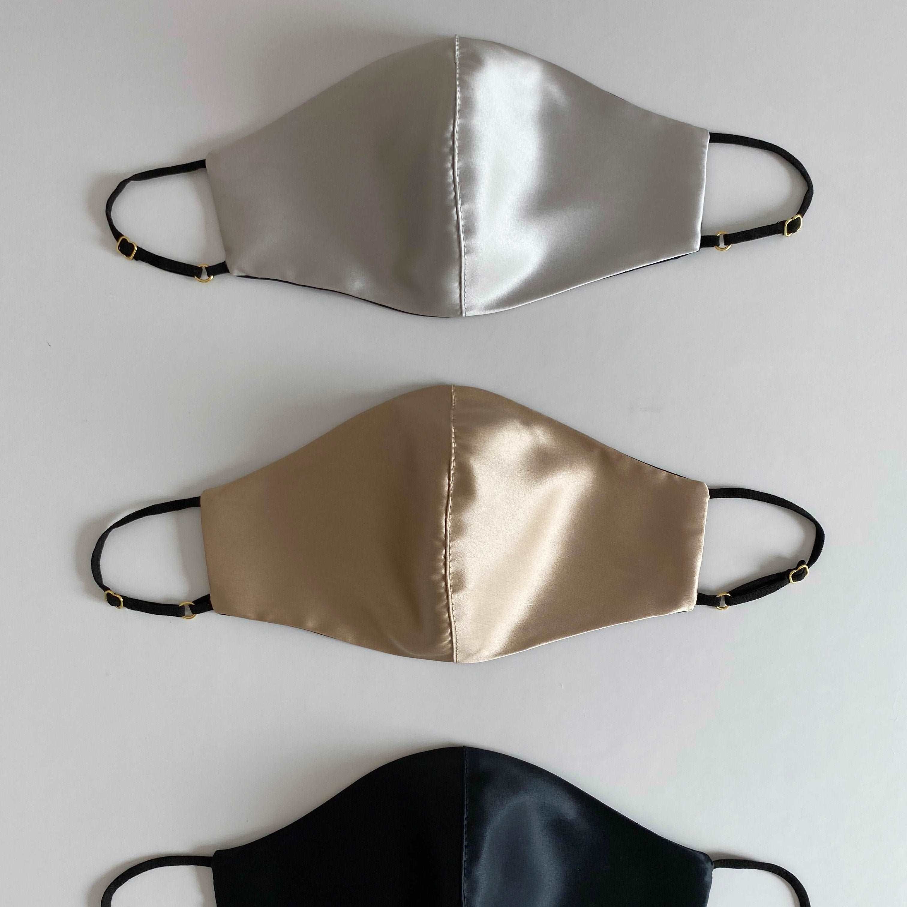 Three silk face masks in French beige, silver, and black, with adjustable straps, displayed side by side against a white background