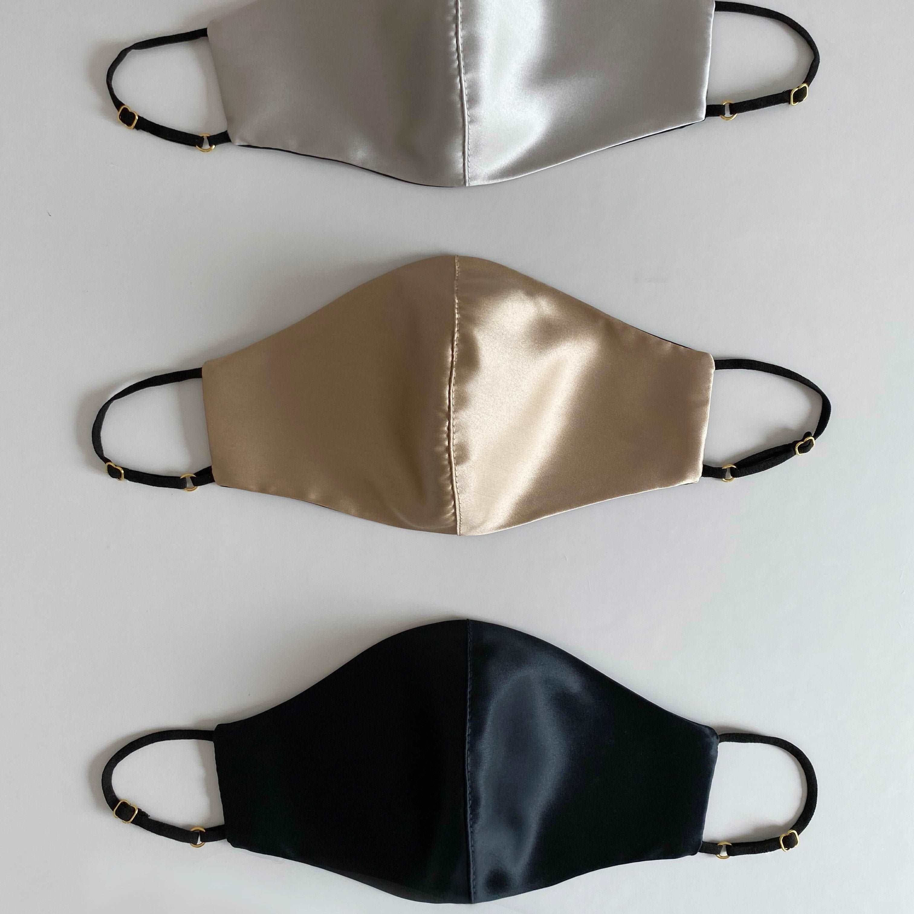 Three silk face masks in French beige, silver, and black, with adjustable straps, displayed side by side against a white background