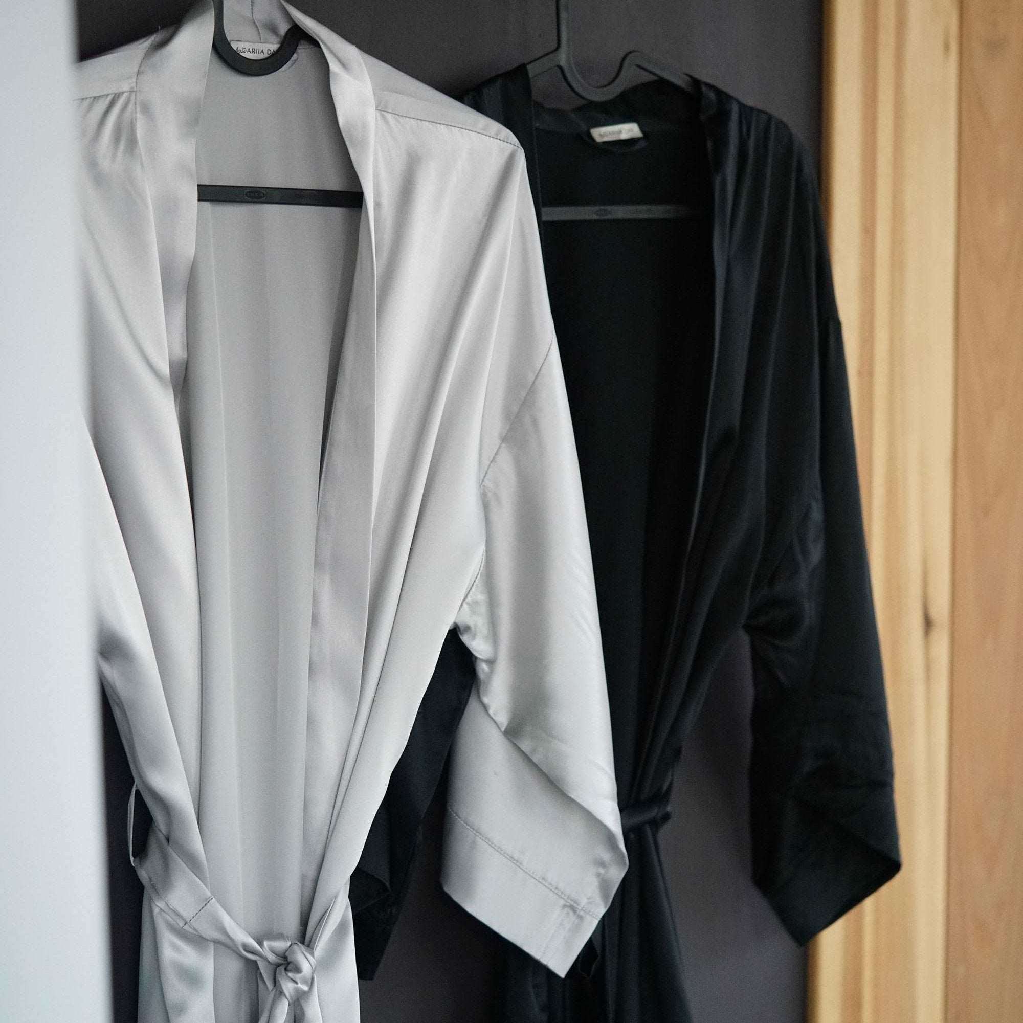 Silver grey and black silk robes by Dariia Day hanging on hooks, showcasing their smooth and elegant fabric.