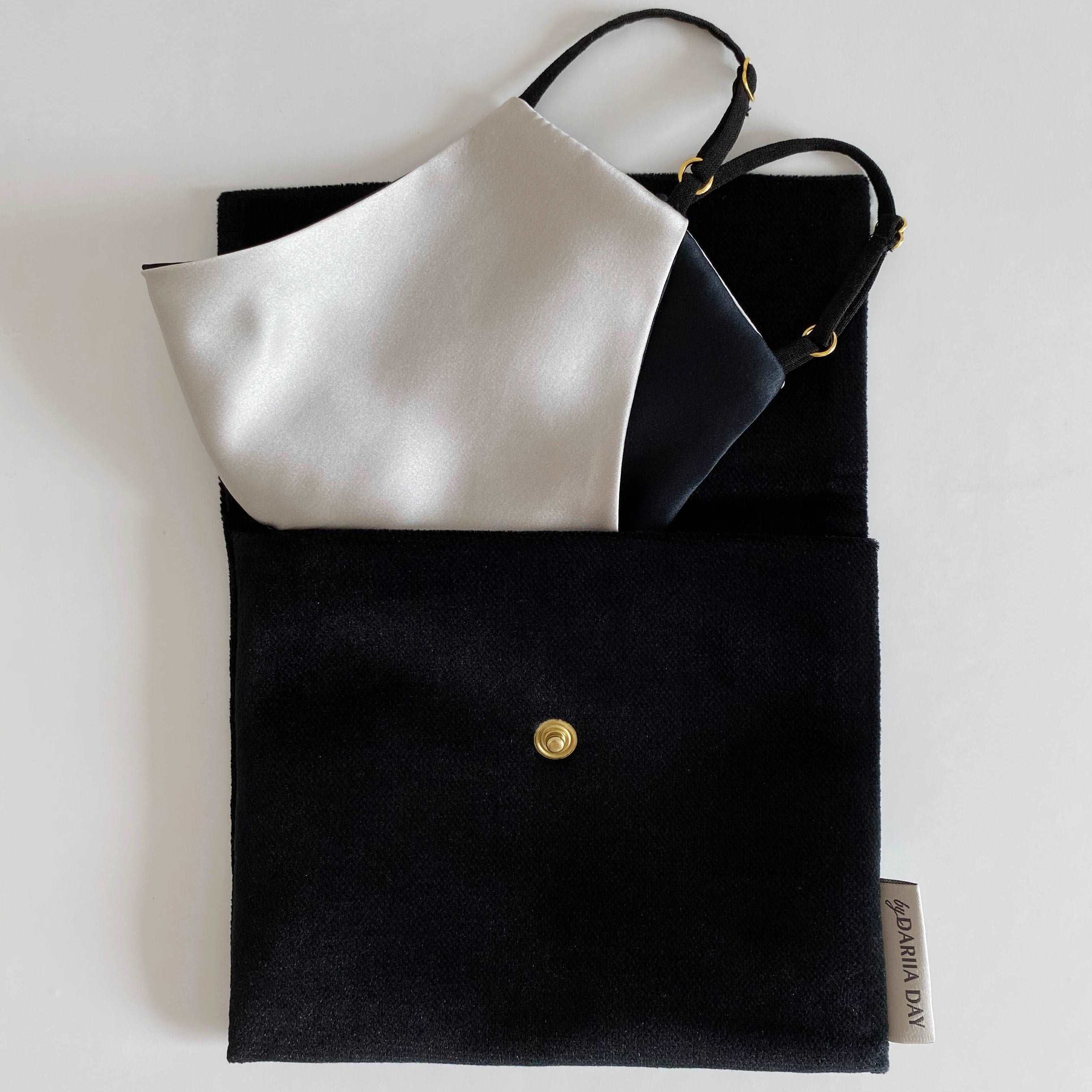 Silver grey silk face mask with black adjustable straps, neatly placed inside a black fabric pouch with a gold button.