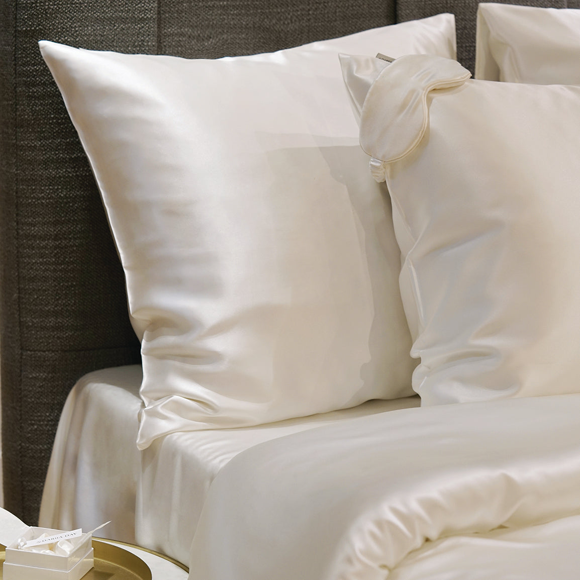 White silk duvet and pillows by Dariia Day, arranged on a bed with a grey headboard and a round bedside table, showcasing the soft and glossy silk texture.