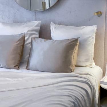 White and silver silk bedding set with a flat sheet and pillows by Dariia Day, displayed on a stylish bed with a bedside table, exuding a luxurious and comfortable vibe.

