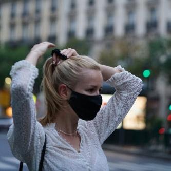 Close-up of a woman adjusting her ponytail while wearing a midnight black silk face mask, with city buildings in the background."