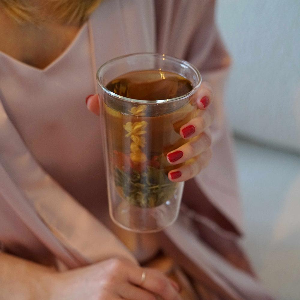 Woman in pink silk robe holding a glass of Morning Blooming Tea by Dariia Day, highlighting the blooming flower inside.