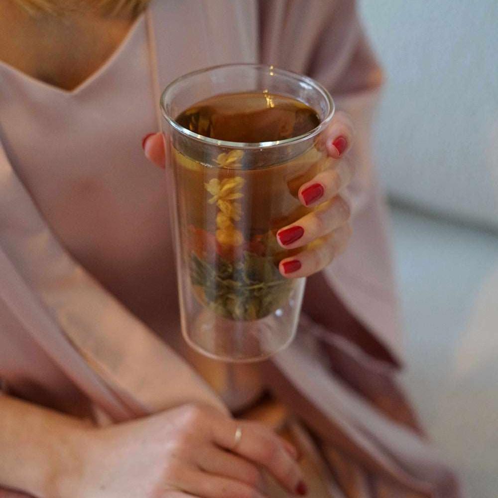 Woman in pink silk robe holding a glass of Morning Blooming Tea by Dariia Day, highlighting the blooming flower inside.