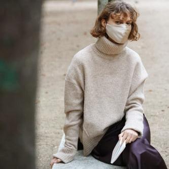 Fashionable woman sitting in a park, wearing a French beige silk face mask and a neutral beige sweater, looking thoughtful.