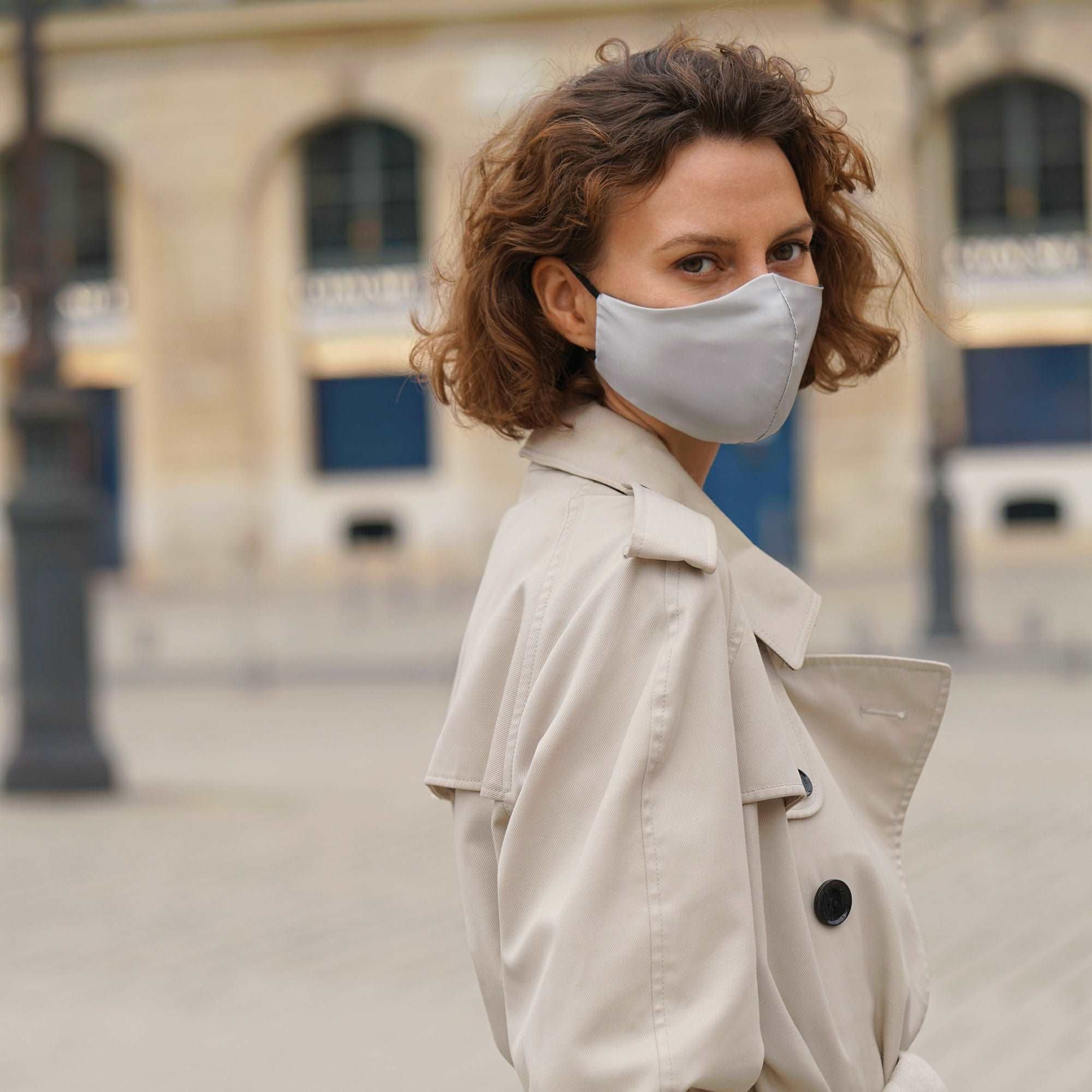 Fashionable woman wearing a silver grey silk face mask with a beige trench coat, posing in an urban setting.