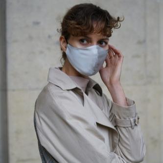 Stylish woman adjusting her silver grey silk face mask while wearing a beige trench coat, standing outdoors.