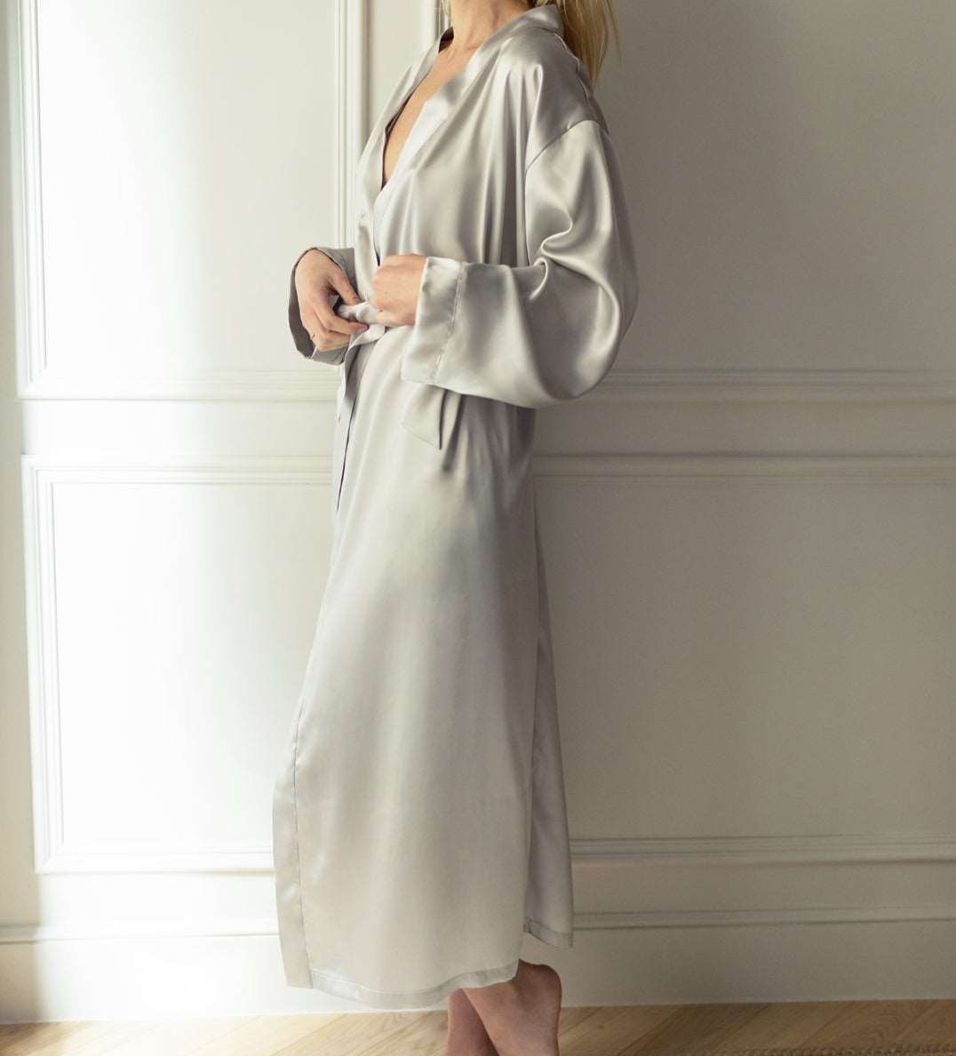 Woman wearing a silver grey silk robe by Dariia Day, standing near a window and tying the belt.